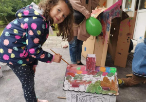 Dziewczynka maluje farbami tekturowy domek.