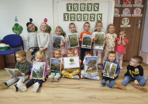 Grupa dzieci stojąca na tle napisu dzień drzewa a dłoniach trzymają ilustracje z różnymi rodzajami drzew