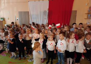 Grupa dzieci w strojach galowych ustawiona na tle flagi biało-czerwonej