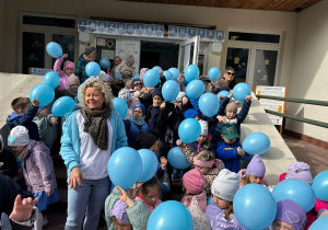 Przedszkolaki z niebieskimi balonami stoją przed wejsciem do przedszkola