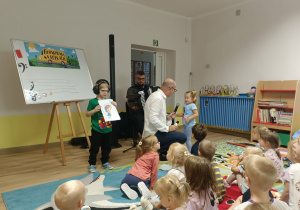 Pan klęczy koło małej dziewczynki obok stoi chłopiec z obrazkiem