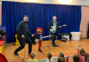Chłopiec przebrany za muzyka gra na gitarze zabawkowej wraz z prowadzącym w akompaniamencie gitarzysty