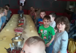 Dzieci stoją przy stole na którym są przygotowane produkty do zrobienia pizzy
