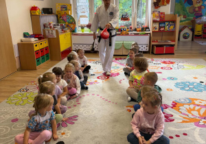 Dzieci siedzą w szeregu dwugrupowym a między nimi pan o taekwondo trzymając w ręku worek treningowy